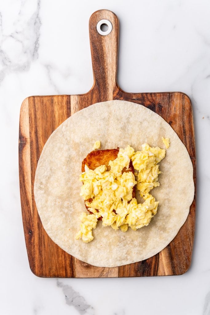 scrambled eggs on a hashbrown patty on a large flour tortilla on a wooden cutting board