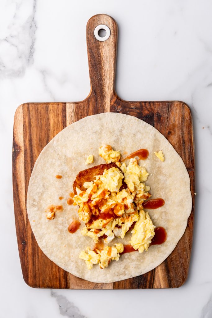 taco sauce topped scrambled eggs and a hashbrown patty on a large flour tortilla on a wooden cutting board