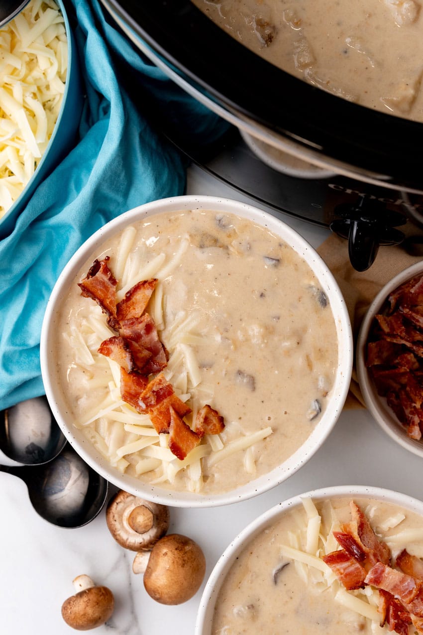 mushroom and potato soup topped with crumbled bacon and shredded swiss cheese in two white bowls