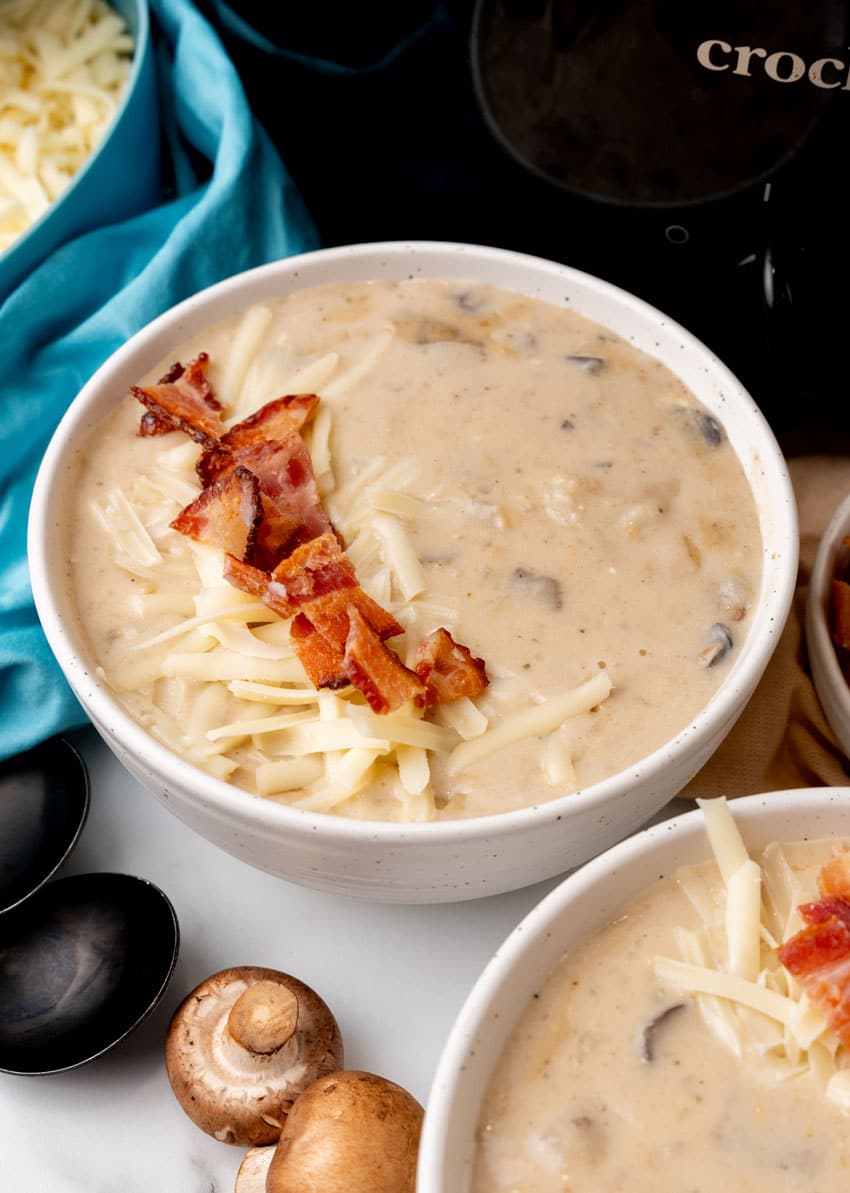 mushroom and potato soup topped with crumbled bacon and shredded swiss cheese in two white bowls