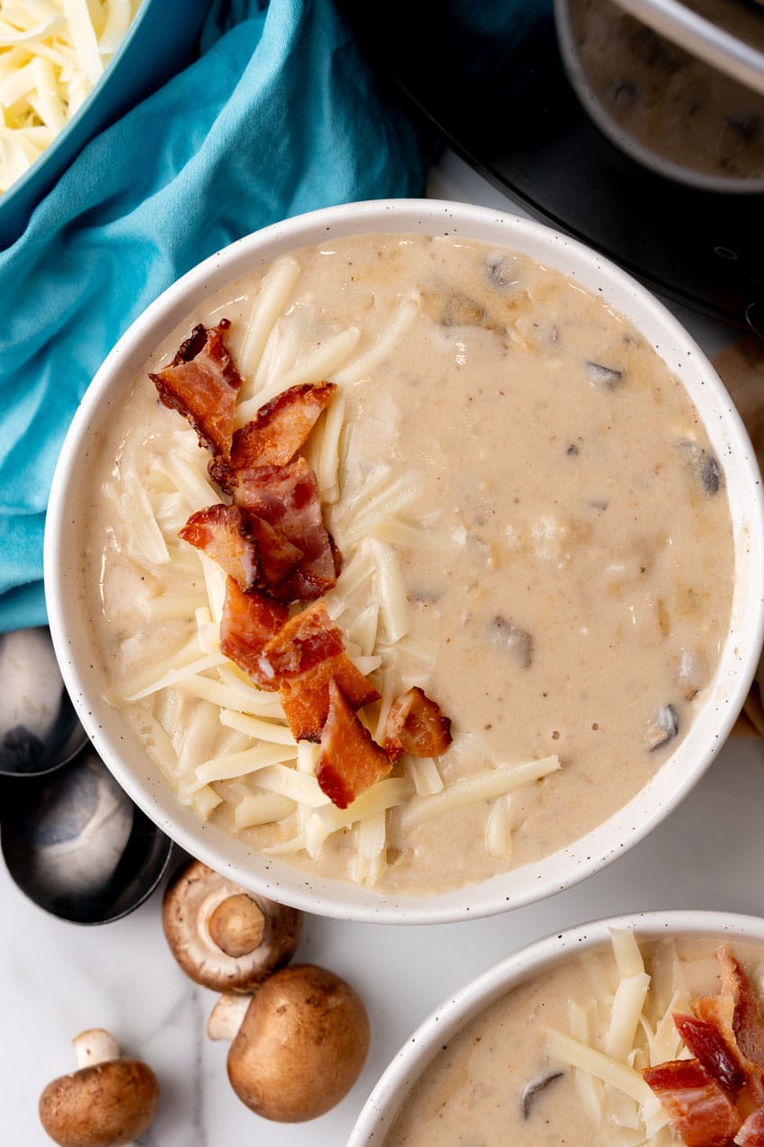 mushroom and potato soup topped with crumbled bacon and shredded swiss cheese in two white bowls