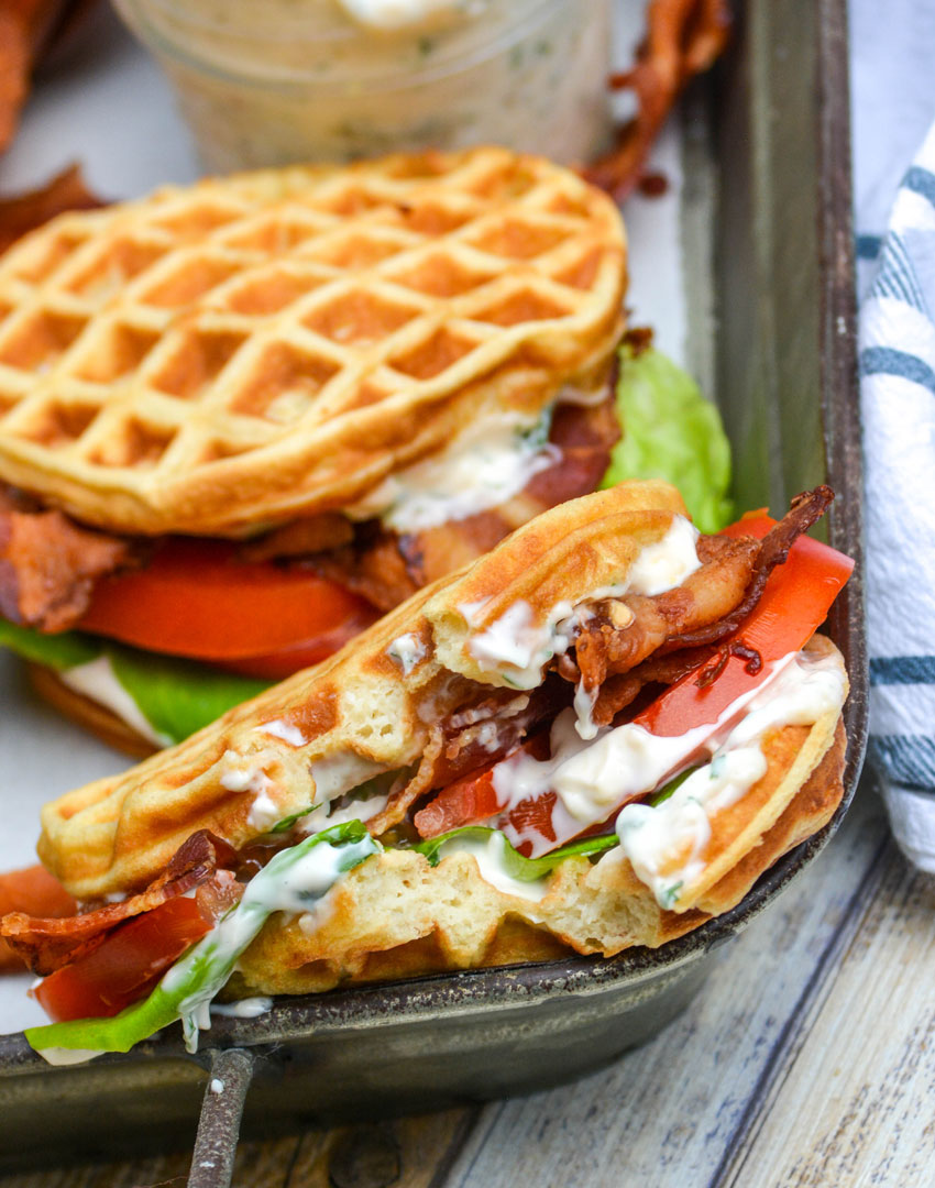 a bacon lettuce and tomato waffle-wich with a bite taken out of the middle resting on the edge of a metal serving tray