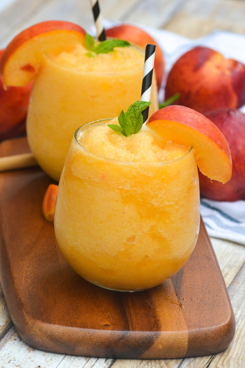two peach wine slushies in glasses on a wooden cutting board surrounded by fresh peaches