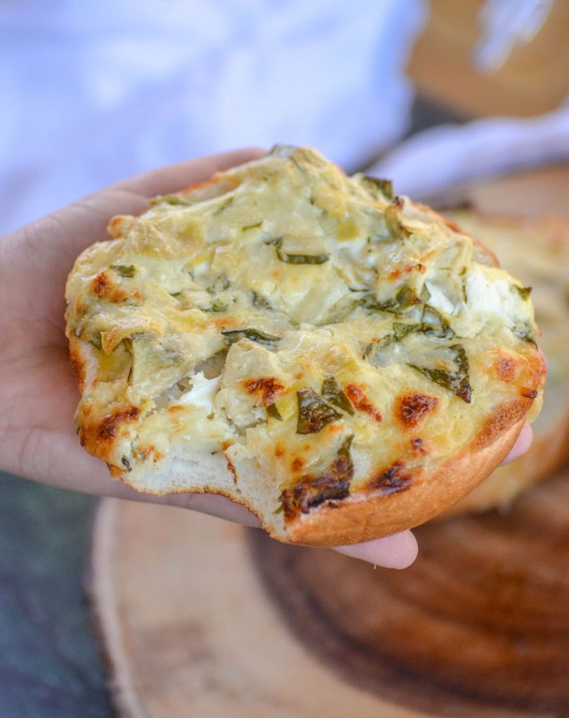 a crispy spinach & artichoke dip covered bagel held up by a hand with a small bite taken out