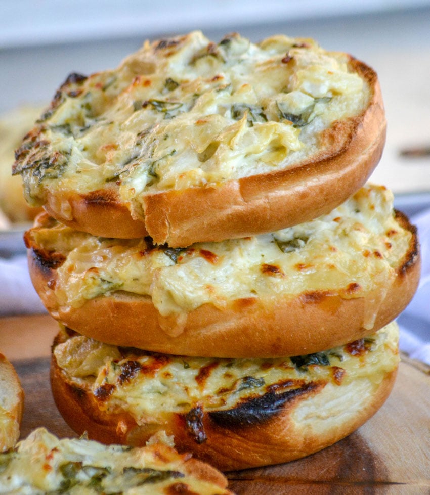 Spinach & Artichoke Bagel Melts stacked three high on a wooden cutting board