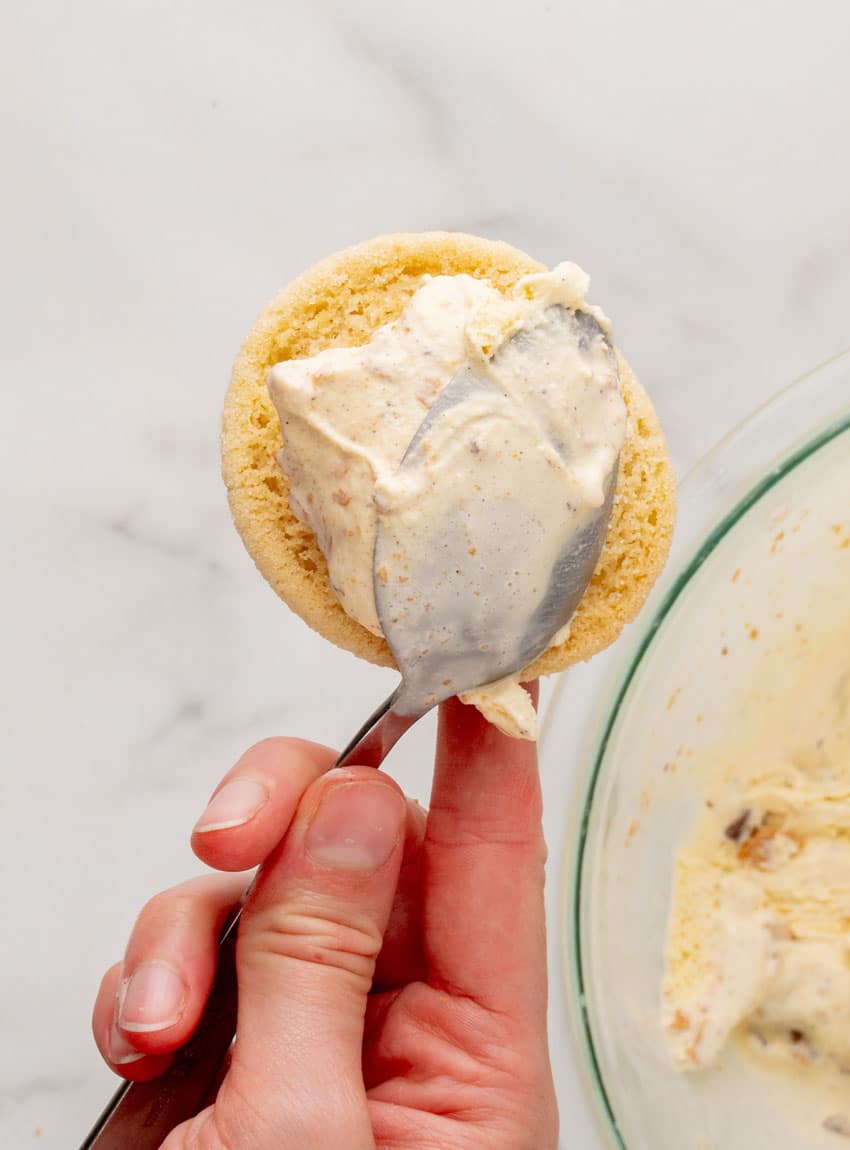 a hand scooping butterfinger flecked vanilla ice cream onto a sugar cookie