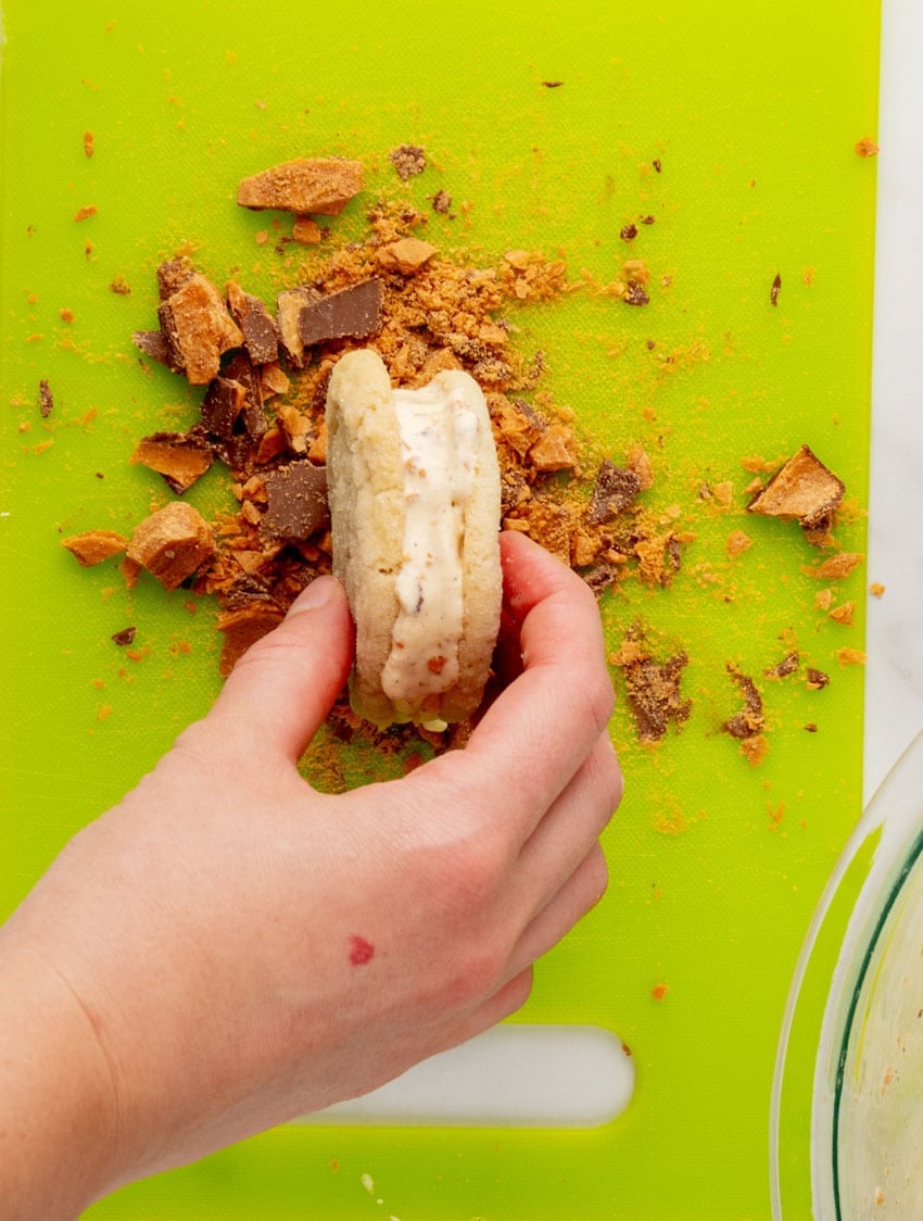 a hand rolling a homemade sugar cookie ice cream sandwich in crushed up butterfinger candy pieces