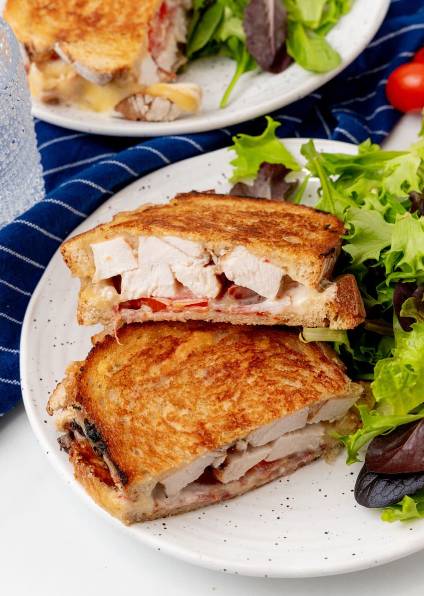 two halves of a chipotle chicken bacon ranch panini on a white plate with leafy greens on the side