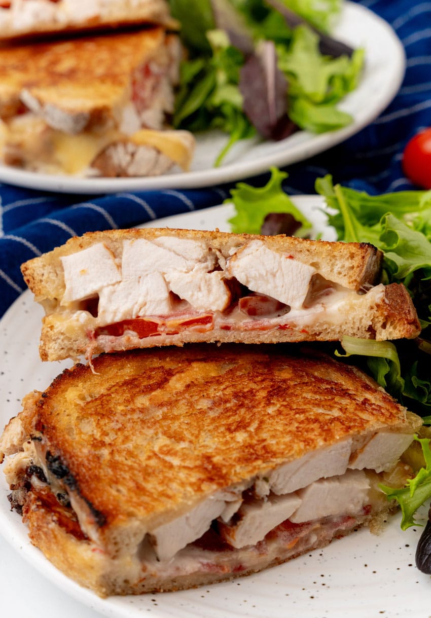 two halves of a chipotle chicken bacon ranch panini on a white plate with leafy greens on the side