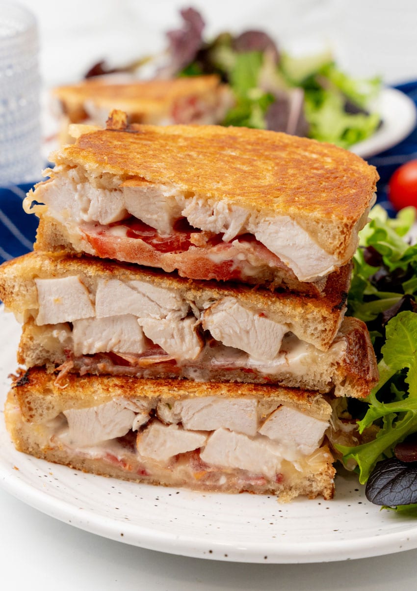 three halves of a chipotle chicken bacon ranch panini sandwich on a white plate with leafy greens on the side