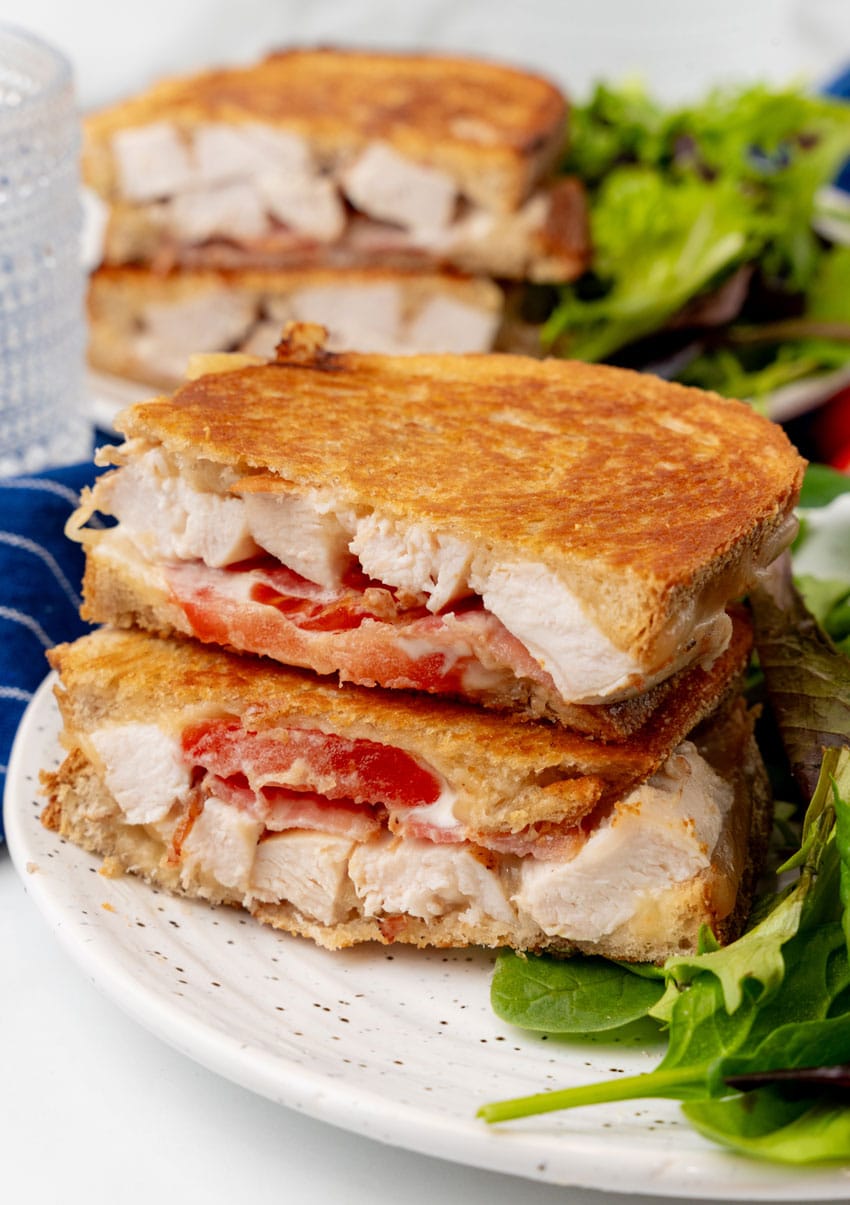 two halves of a chipotle chicken bacon ranch panini on a white plate with leafy greens on the side