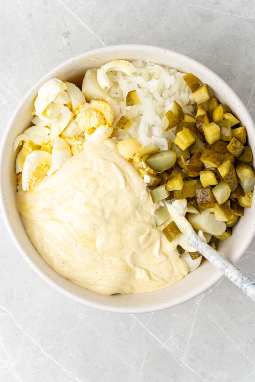 dill pickle potato salad ingredients in a large white mixing bowl with a white spoon resting on the side