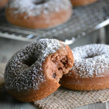 a bitten baked chocolate malt donut resting on another donut on a piece of burlap on a green table