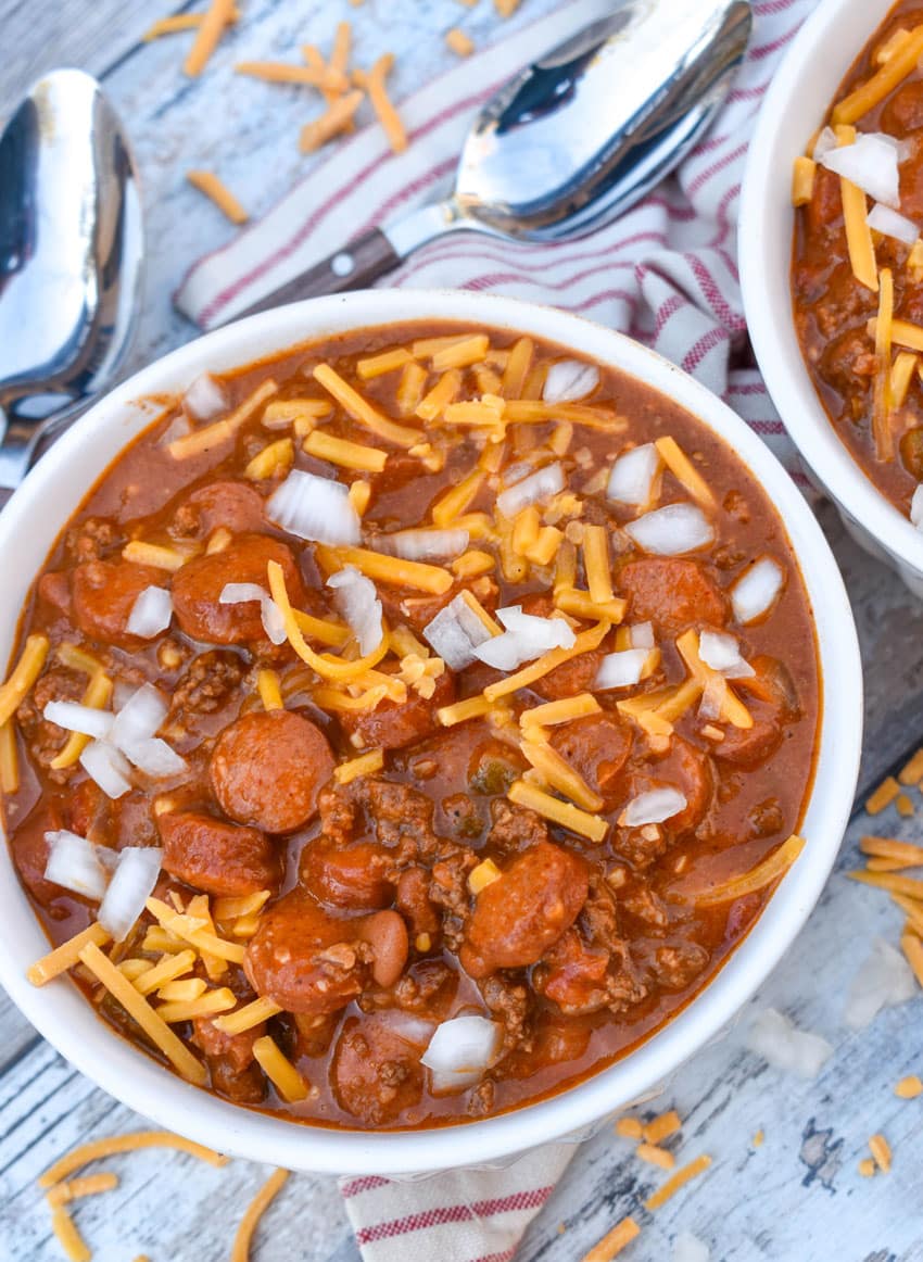 slow cooker chili cheese dog chili topped with shredded cheddar cheese and diced white onion in two white bowls