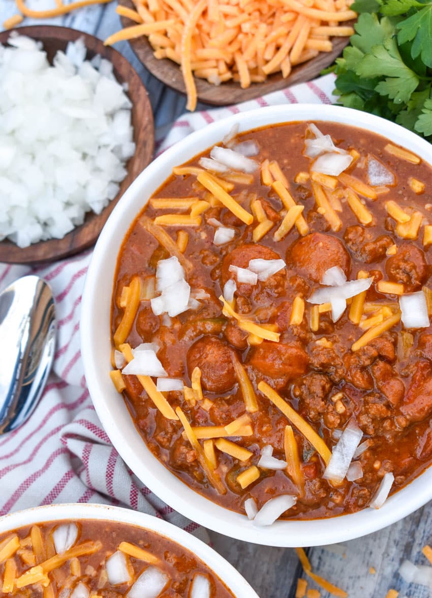 slow cooker chili cheese dog chili topped with shredded cheddar cheese and diced white onion in two white bowls