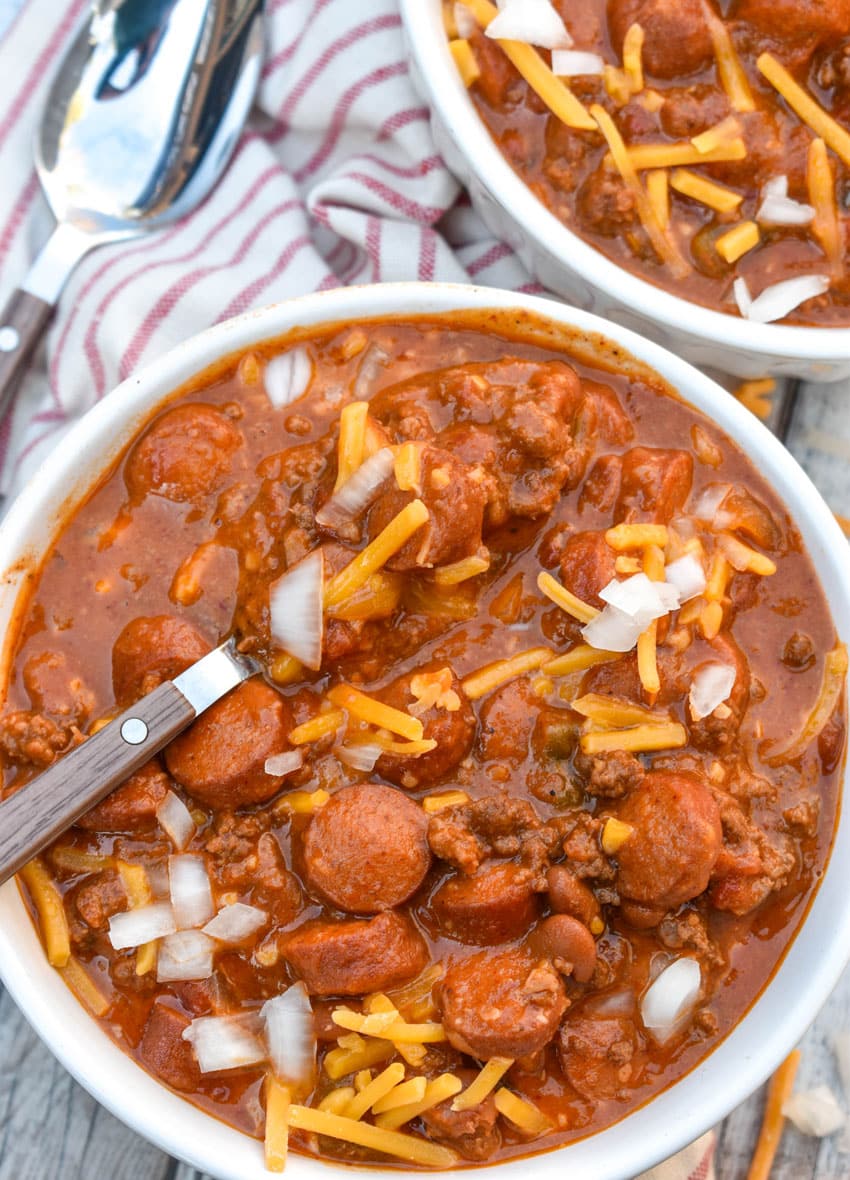 slow cooker chili cheese dog chili topped with shredded cheddar cheese and diced white onion in two white bowls