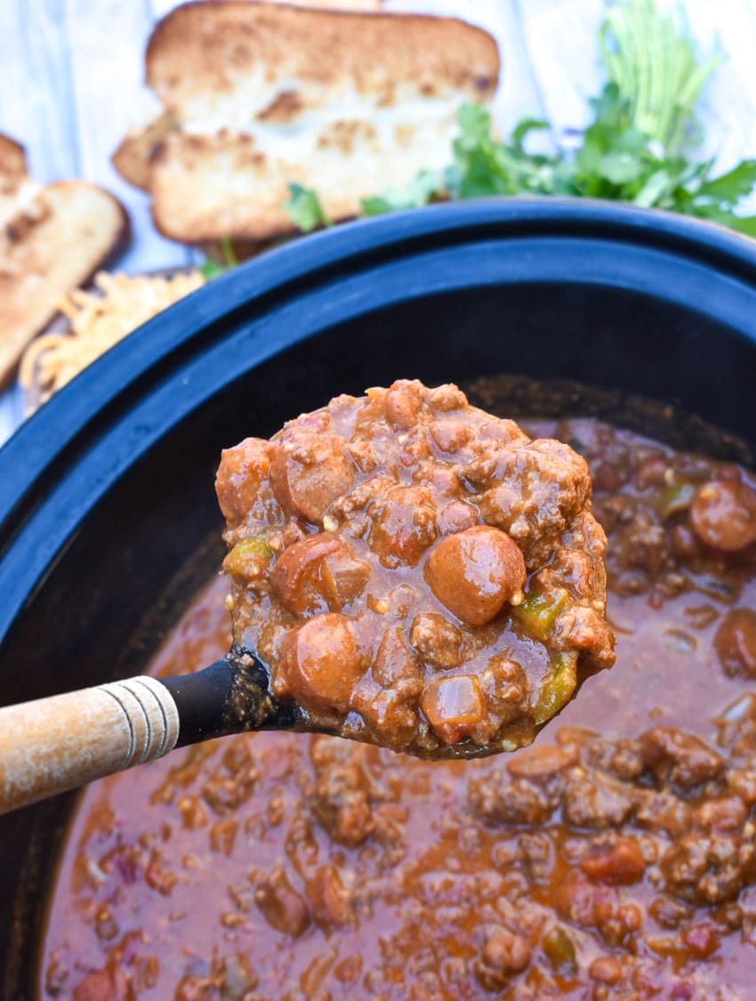 a wooden ladle scooping homemade chili cheese dog chili out of a black crockpot