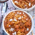 slow cooker chili cheese dog chili topped with shredded cheddar cheese and diced white onion in two white bowls