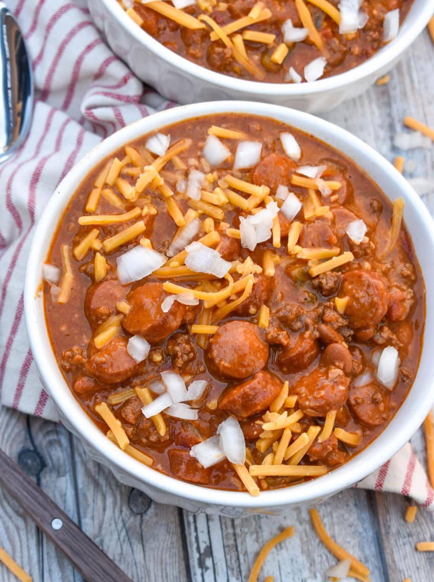 slow cooker chili cheese dog chili topped with shredded cheddar cheese and diced white onion in two white bowls