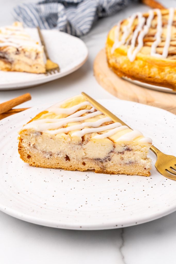 a slice of cinnamon bun cheesecake on a white plate with a gold fork on the side