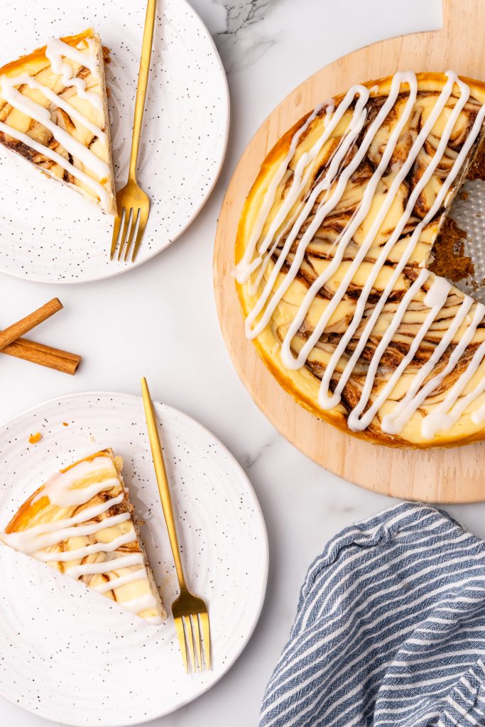 slices of cinnamon bun cheesecake on a white plates with gold forks on the side