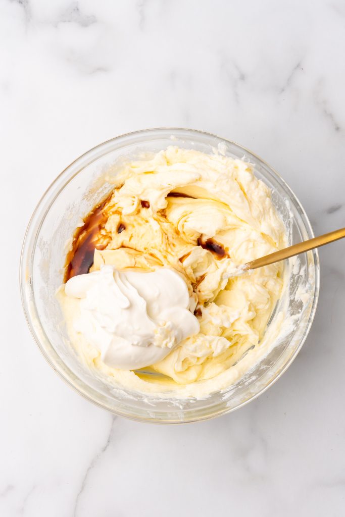 sour cream and vanilla extract being added to a bowl of whipped cream cheese and sugar