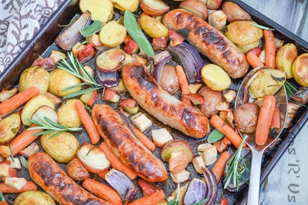 Sausage, Apple, And Herb Sheet Pan Supper shown on a dark brown sheet pan with fresh herbs and a silver spoon for serving