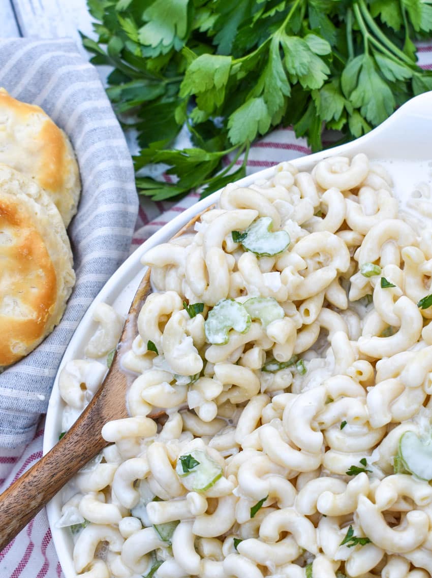 a wooden spoon scooping homemade creamy macaroni salad out of a white serving bowl