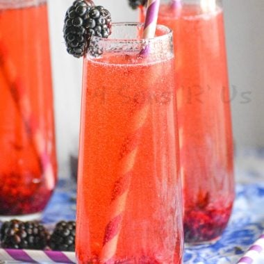 THREE STEMLESS WINE FLUTES FILLED WITH BLACKBERRY LAVENDAR CHAMPAGNE COCKTAIL WITH PAPER STRAWS AND A FRESH BLACKBERRY ON THE RIM FOR GARNISH