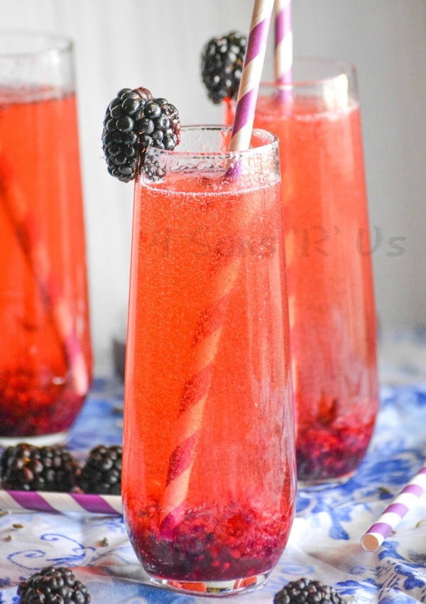 THREE STEMLESS WINE FLUTES FILLED WITH BLACKBERRY LAVENDAR CHAMPAGNE COCKTAIL WITH PAPER STRAWS AND A FRESH BLACKBERRY ON THE RIM FOR GARNISH