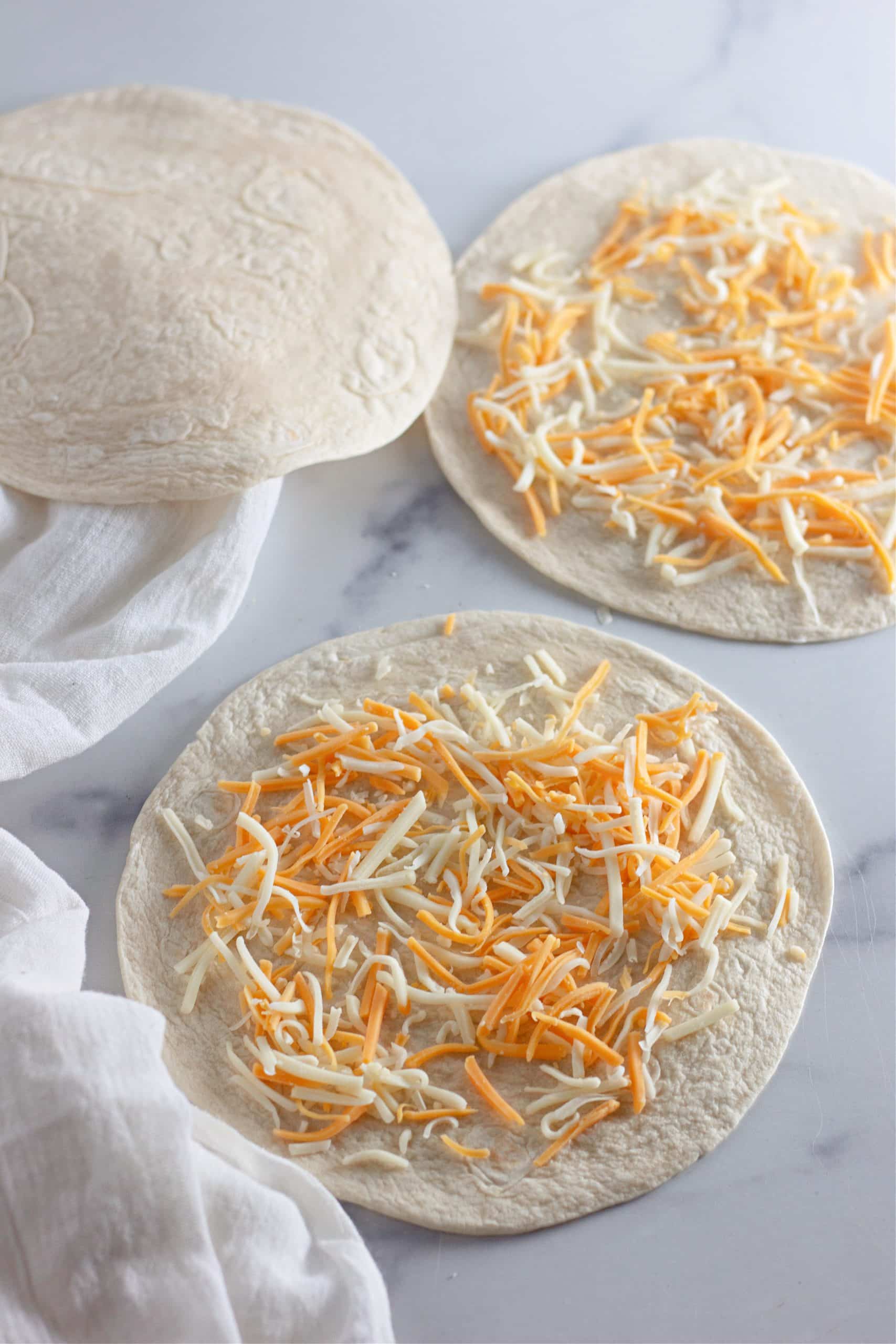shredded cheese on flour tortillas on a marble counter top