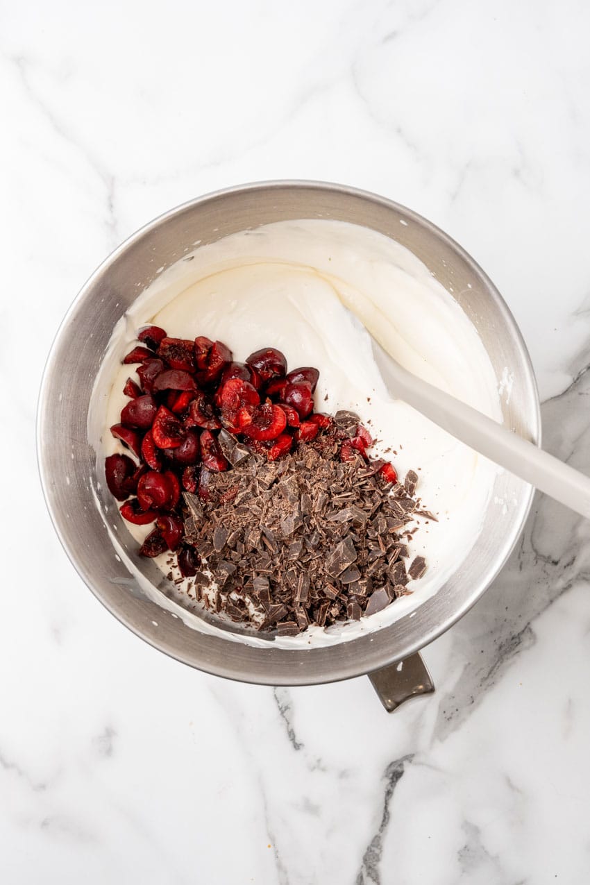 chopped sweet dark cherries and chocolate shavings being mixed into whipped cream in the bowl of a stand mixer