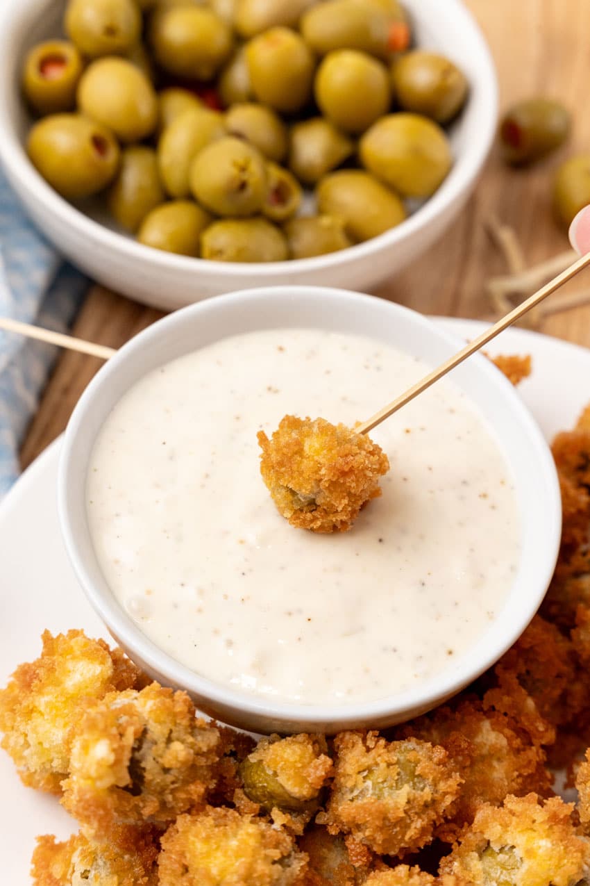 a fried olive on a wooden toothpick being dipped into a small bowl of garlic aioli