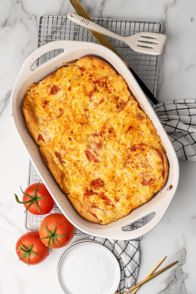 baked cheese topped tomato biscuit casserole in a white casserole dish next to ripe red tomatoes