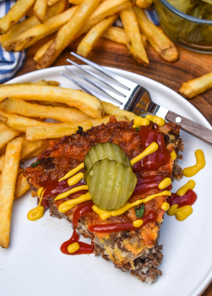 a slice of cheeseburger pie topped with ketchup and mustard next to crispy french fries on a white plate