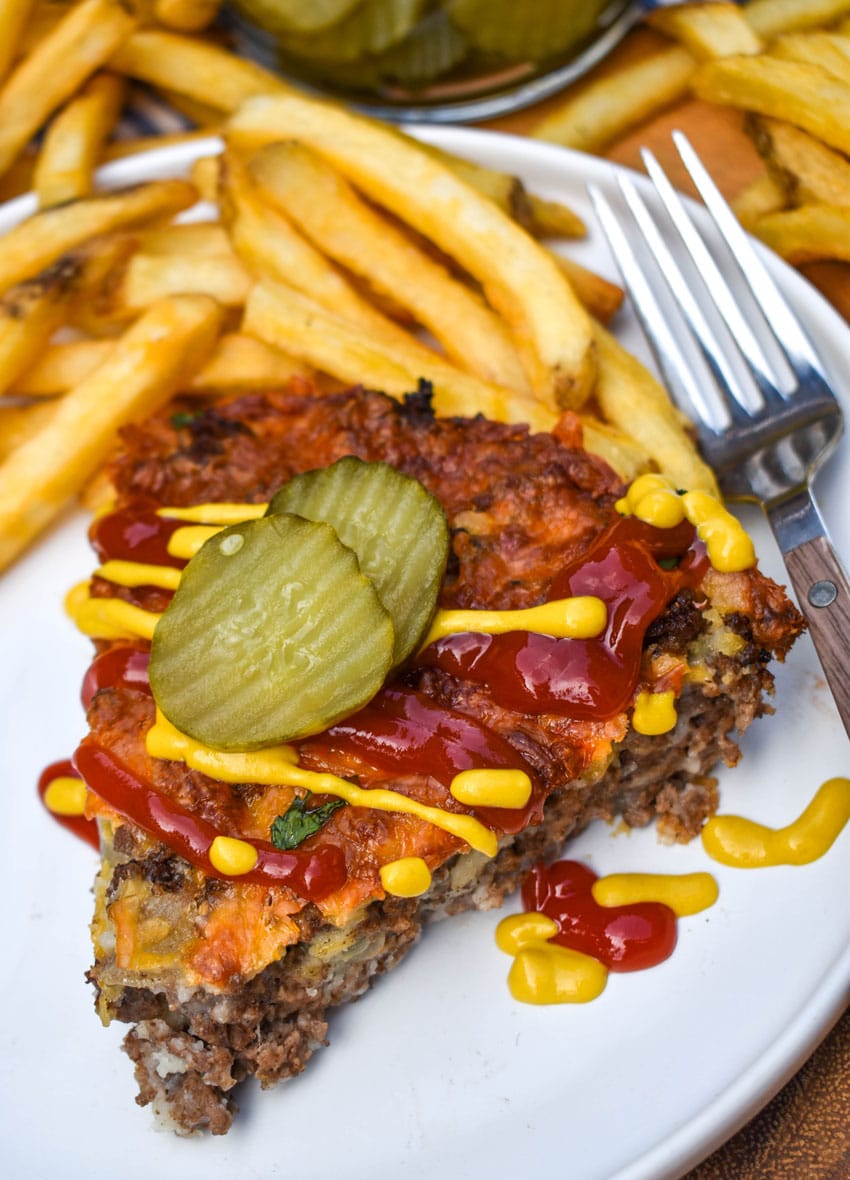 a slice of cheeseburger pie topped with ketchup and mustard next to crispy french fries on a white plate
