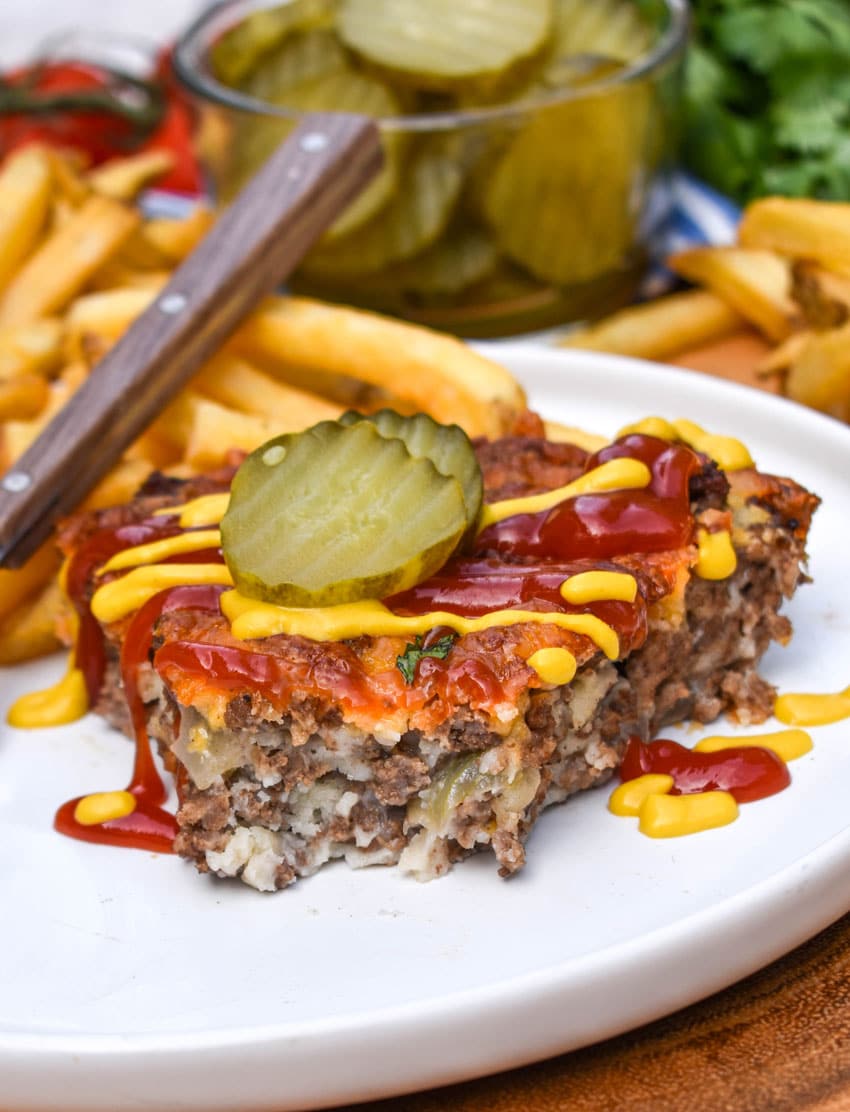 a slice of cheeseburger pie topped with ketchup and mustard next to crispy french fries on a white plate
