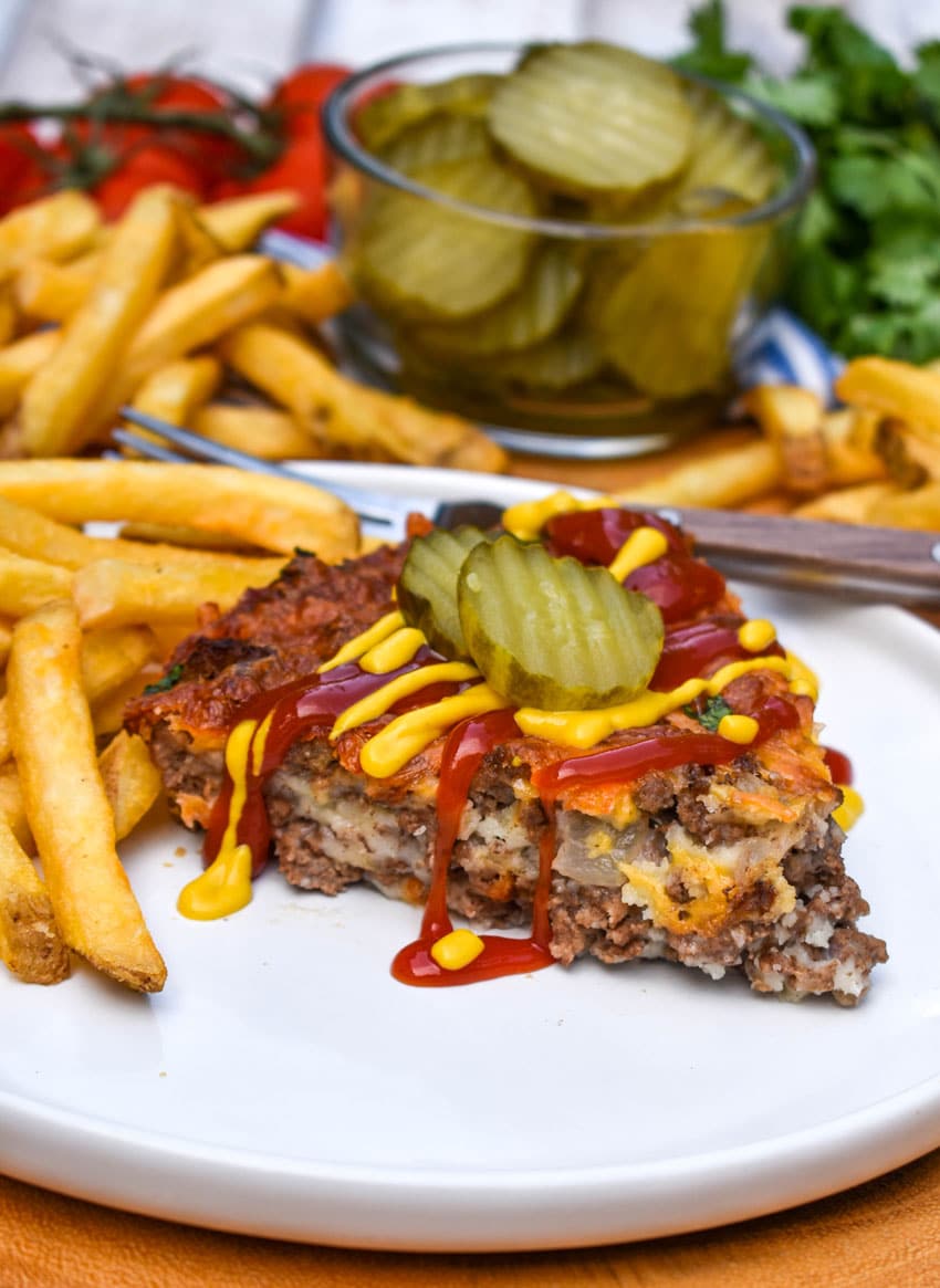 a slice of cheeseburger pie topped with ketchup and mustard next to crispy french fries on a white plate