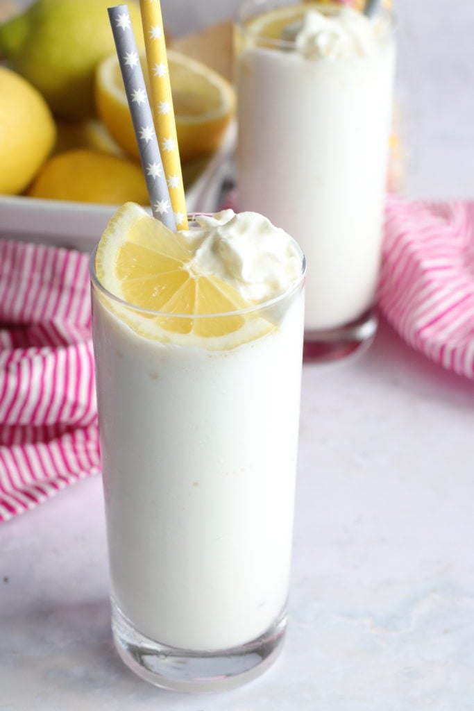 copy cat chick fil a frosted lemonade served in a slender glass and topped with whipped cream, a slice of lemon and a gray and yellow paper straw