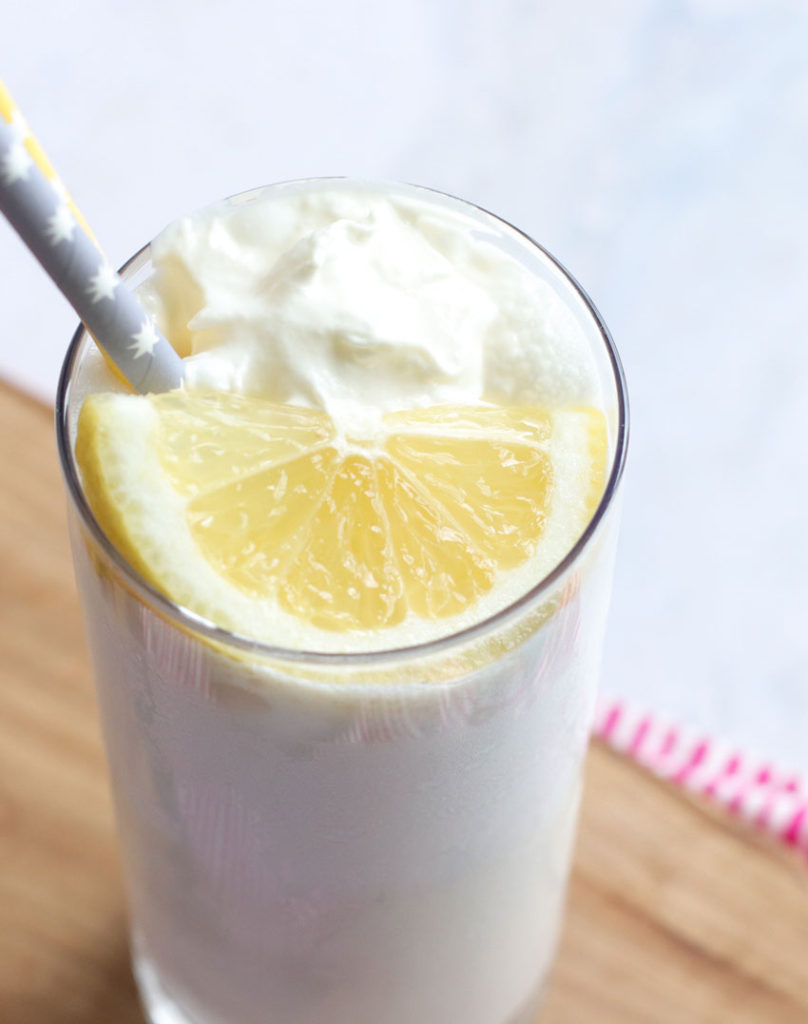 copy cat chick fil a frosted lemonade served in a slender glass and topped with whipped cream, a slice of lemon and a gray paper straw