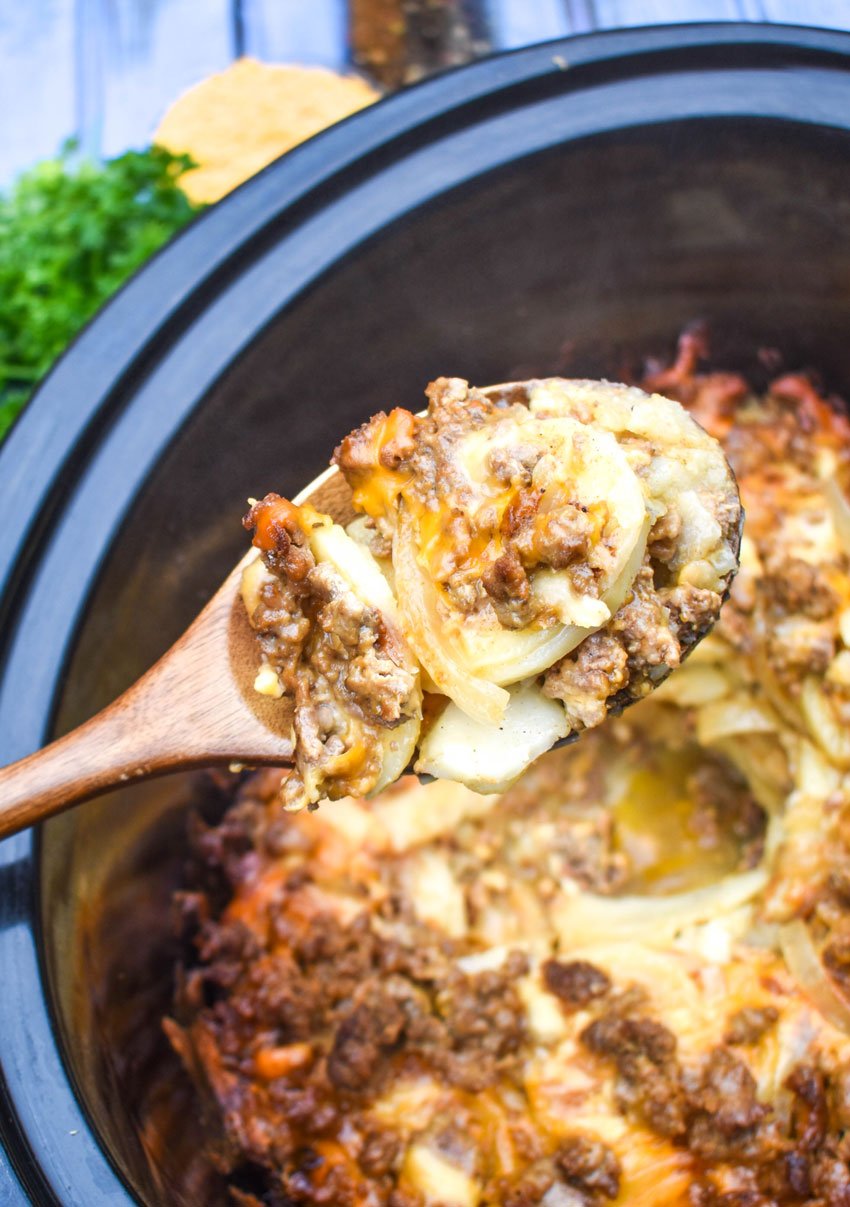 a wooden spoon holding a scoop of cheesy ground beef and potatoes above a black slow cooker