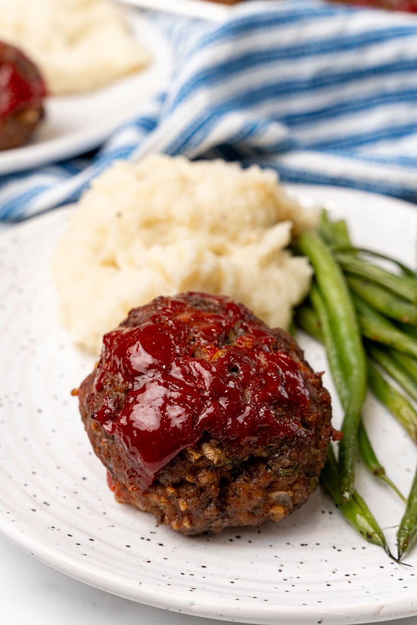 a small meatloaf next to a scoop of mashed potatoes and roasted green beans on a white plate