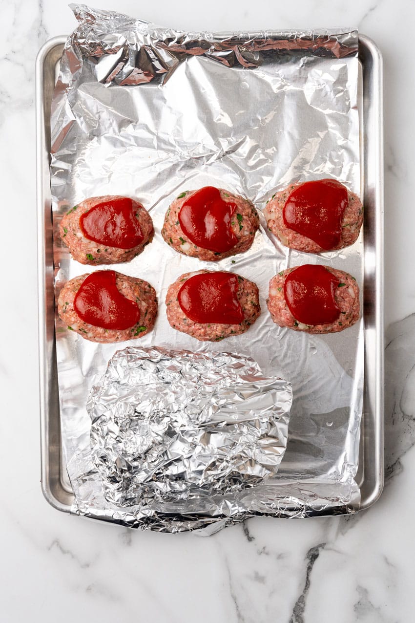 ketchup topped unbaked mini meatloaves on a metal sheet pan next to a aluminum foil packet