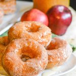 glazed apple cider donuts stacked in a pile on a white plate with pooling glaze around the edges