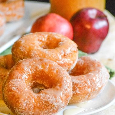 glazed apple cider donuts stacked in a pile on a white plate with pooling glaze around the edges