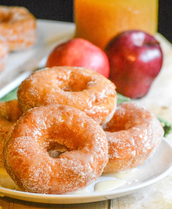 glazed apple cider donuts stacked in a pile on a white plate with pooling glaze around the edges