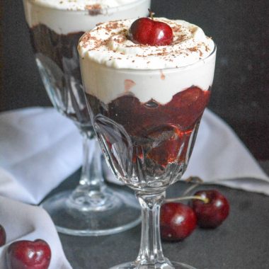 TWO BLACK FOREST CHERRY CHEESECAKE DESSERT CUPS WITH A GRAY CLOTH NAPKIN IN THE BACKGROUND AND FRESH CHERRIES ON THE SIDE