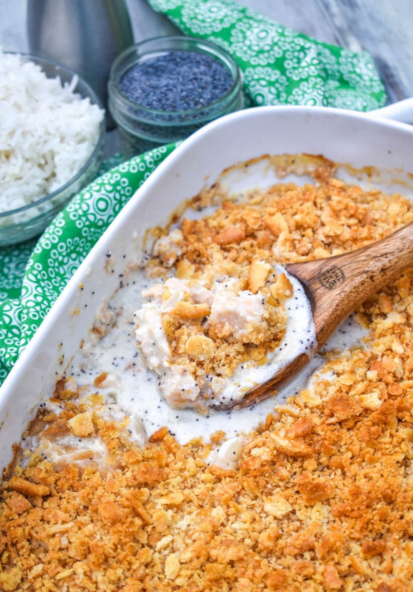 a wooden spoon scooping creamy chicken casserole with poppy seeds out of a white baking dish