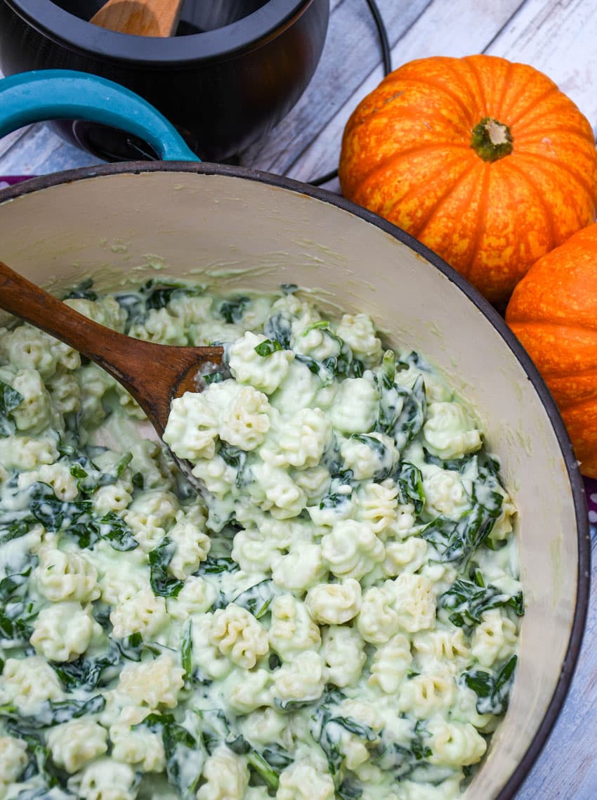 a wooden spoon scooping white cheddar mac and cheese with spinach out of a large cast iron pot