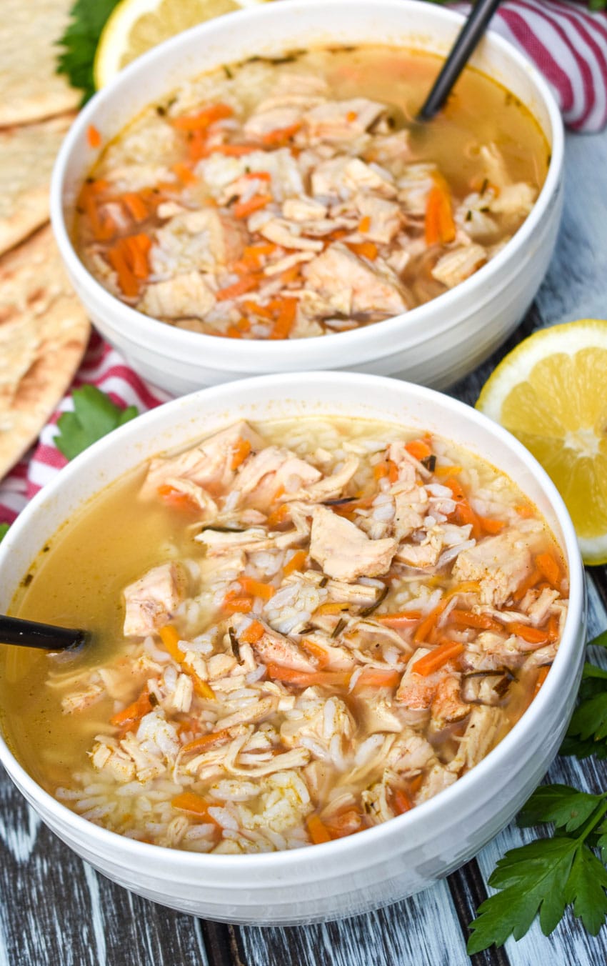 greek lemon chicken soup with rice in two white bowls with sliced lemons and fresh herbs on the side